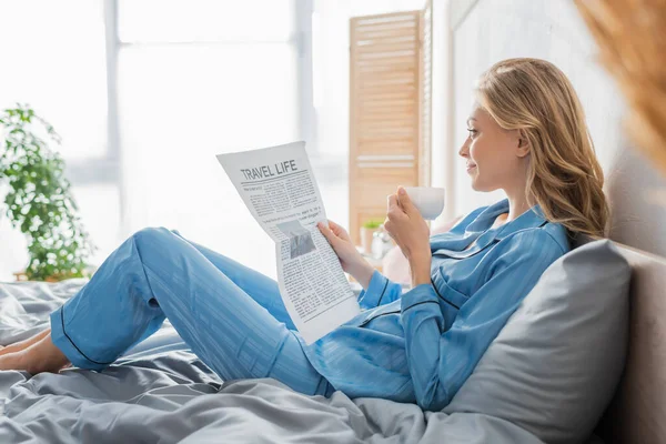 Vista lateral de la joven despreocupada leyendo el periódico de viaje y sosteniendo la taza de café en la cama - foto de stock