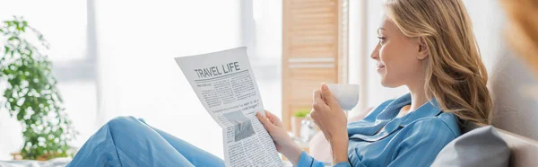 Вид сбоку на молодую женщину, читающую газету о путешествиях и держащую чашку кофе в спальне, баннер — стоковое фото