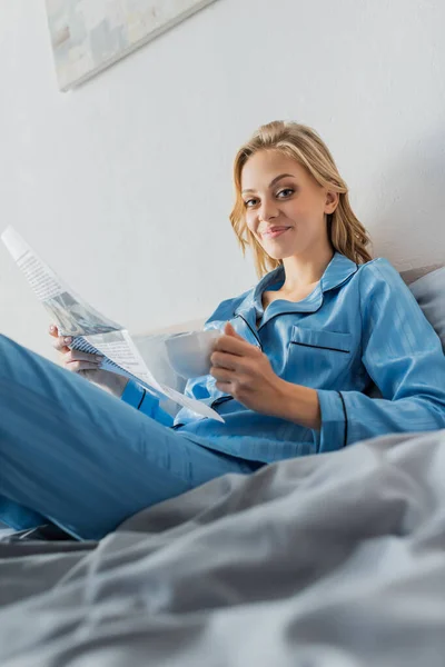 Happy young woman holding newspaper and cup of coffee while resting in bed — Stock Photo