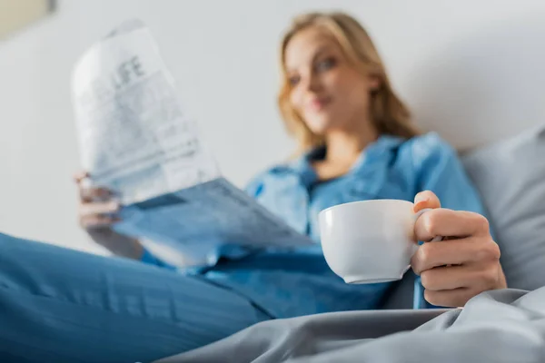 Cup of coffee in hand of young woman with newspaper on blurred background — Stock Photo
