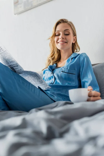 Happy young woman holding newspaper and looking at cup of coffee while resting in bed — Stock Photo