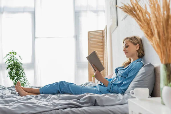 In voller Länge erfreut junge Frau liest Buch in der Nähe Tasse Kaffee auf dem Nachttisch — Stockfoto