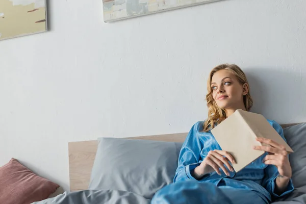 Стомлена молода жінка в блакитному шовковому нічному одязі тримає книгу в спальні — стокове фото