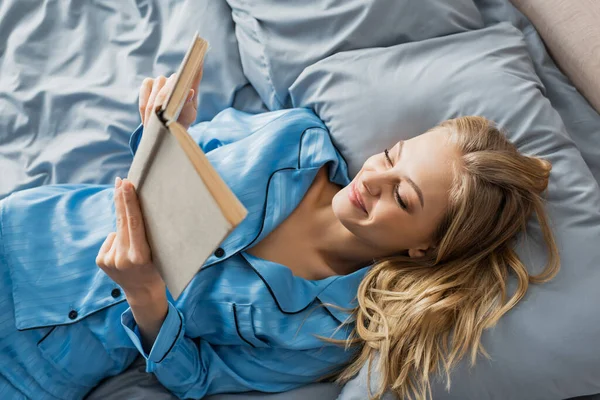 Вид сверху довольной молодой женщины в синей шелковой ночной одежде, читающей книгу на кровати — стоковое фото