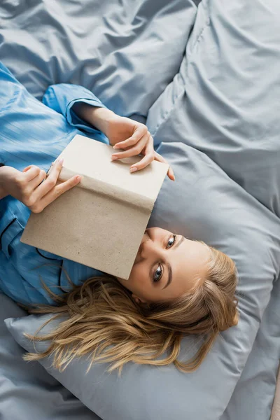 Vista dall'alto di compiaciuta giovane donna in camicia da notte di seta blu che copre il viso con libro a letto — Foto stock
