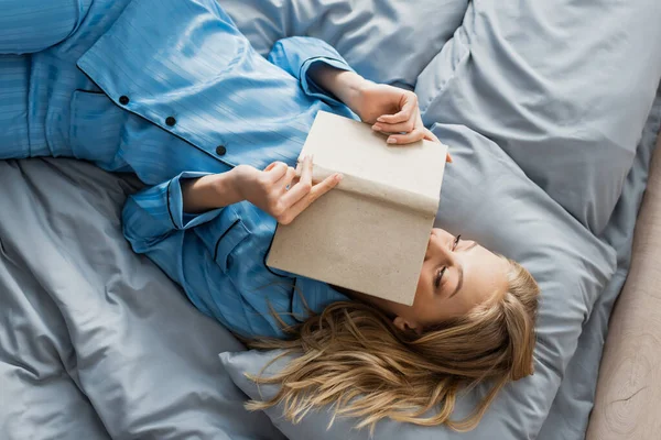 Vista superior de mujer joven satisfecha en la cara de la cubierta de la ropa de dormir de seda azul con el libro en la cama - foto de stock