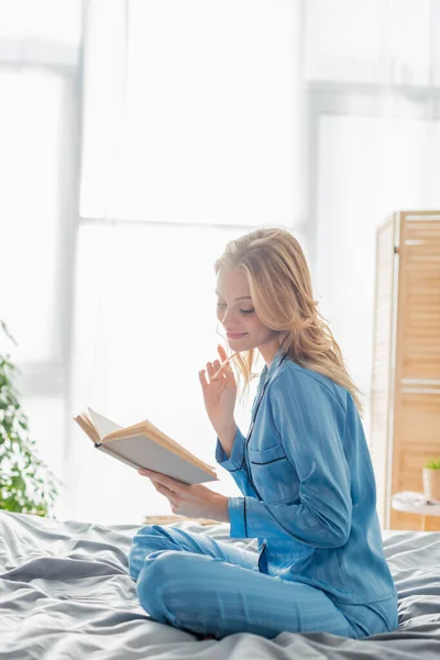 Satisfied young woman in blue silk nightwear reading book while resting on weekend — Stock Photo