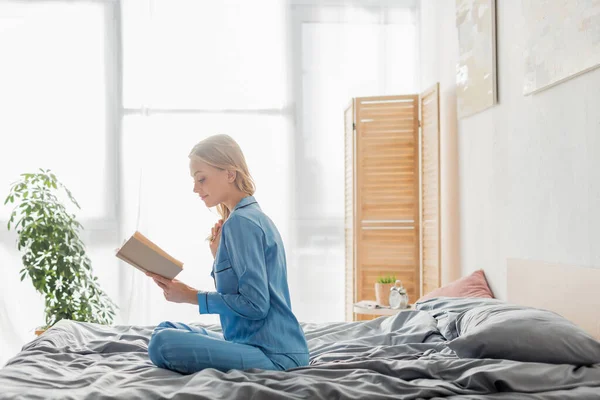 Jovem mulher despreocupada em roupa de seda azul leitura livro no apartamento moderno — Fotografia de Stock