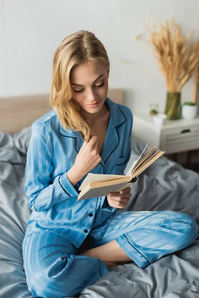 Blonde jeune femme en soie bleue livre de lecture de vêtements de nuit tout en se reposant le week-end — Photo de stock