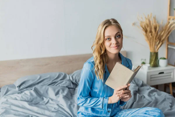 Задоволена жінка в блакитному шовковому нічному одязі тримає книгу під час відпочинку на вихідних — стокове фото