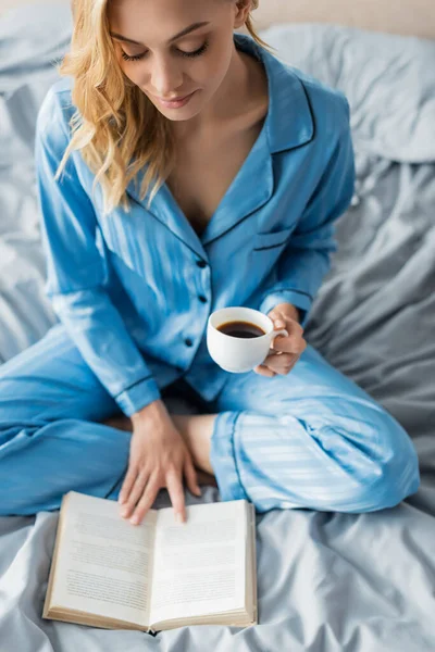 Overhead view of young woman in blue silk nightwear reading book while holding cup of coffee — Stock Photo