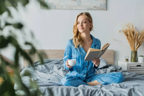 Красива молода жінка в блакитному шовковому нічному одязі тримає книгу і чашку кави в сучасній спальні — стокове фото