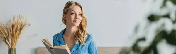 Bella giovane donna in camicia da notte di seta blu che tiene libro mentre guarda lontano a casa, banner — Foto stock