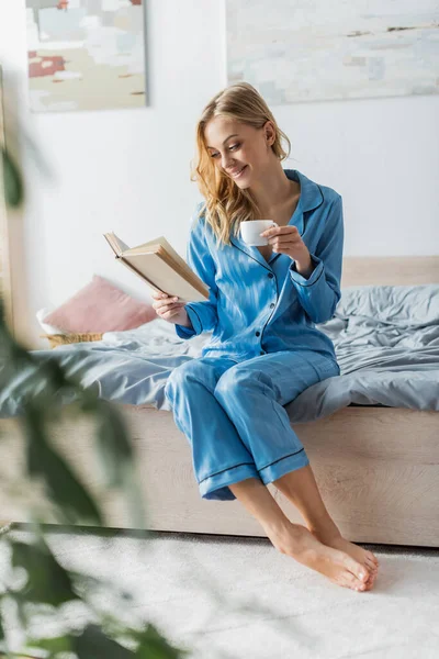 Mulher sorrindo no livro de leitura de roupa de seda azul nightwear enquanto segurando xícara de café no quarto — Fotografia de Stock