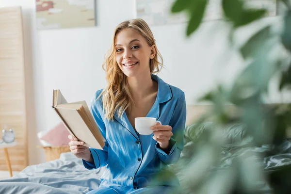 Charming woman in blue silk nightwear reading book and holding cup of coffee in bedroom — Stock Photo