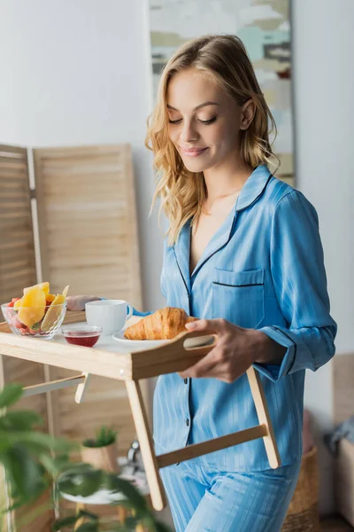 Zufriedene junge Frau im blauen Pyjama mit Tablett und Frühstück im Schlafzimmer — Stockfoto