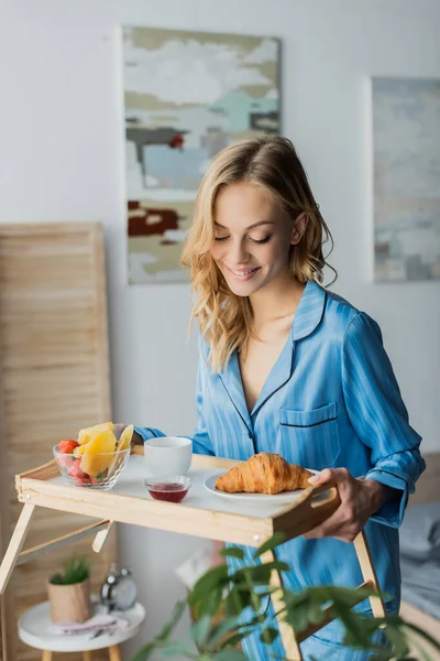 Mujer feliz en pijama azul mirando bandeja con desayuno en el dormitorio - foto de stock