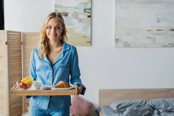 Alegre mujer en azul pijama celebración bandeja con desayuno cerca de la cama en apartamento moderno - foto de stock