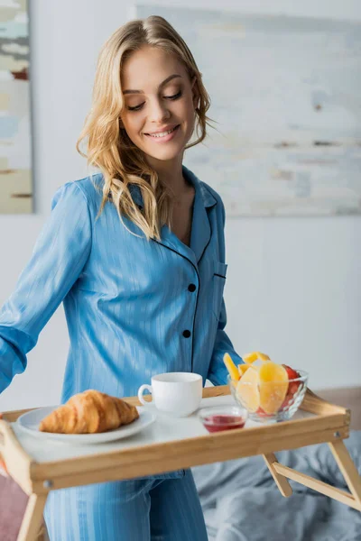 Улыбающаяся молодая женщина в синей ночной рубашке держит поднос с завтраком в спальне — стоковое фото