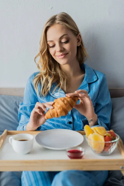 Улыбающаяся молодая женщина в синей пижаме держит свежий круассан возле подноса во время завтрака в постели — стоковое фото