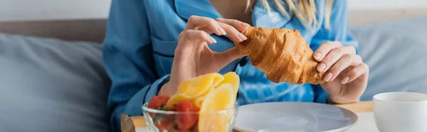 Vista cortada da jovem mulher em pijama azul segurando croissant fresco perto da bandeja enquanto toma café da manhã na cama, banner — Fotografia de Stock