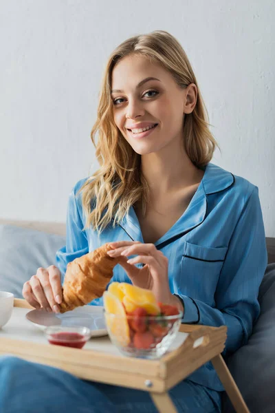 Jolie femme en pyjama bleu tenant croissant frais près du plateau tout en prenant le petit déjeuner au lit — Photo de stock