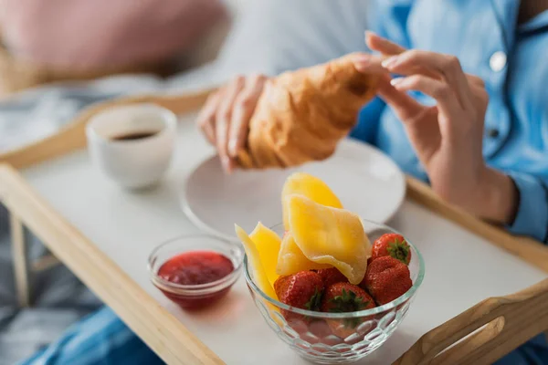 Vista cortada da mulher segurando croissant fresco perto da bandeja com geléia e manga seca enquanto toma café da manhã na cama — Fotografia de Stock