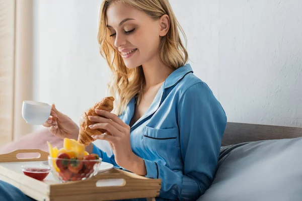 Sonriente joven en pijama sosteniendo una taza de café y croissant cerca de la bandeja mientras desayuna en la cama - foto de stock