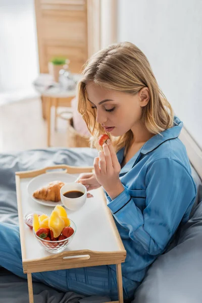 Rubia joven en pijama sosteniendo taza de café y fresa fresca cerca de bandeja mientras desayuna en la cama - foto de stock
