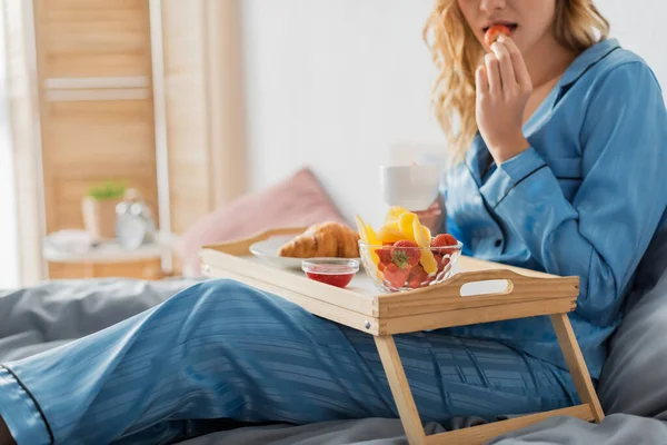 Vue recadrée de la femme tenant une tasse de café et mangeant fraise fraîche près du plateau tout en prenant le petit déjeuner au lit — Photo de stock