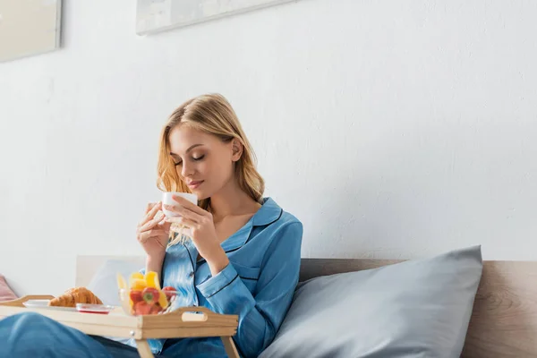 Bonita mujer sosteniendo la taza mientras disfruta del sabor del café cerca de la bandeja del desayuno en la cama - foto de stock