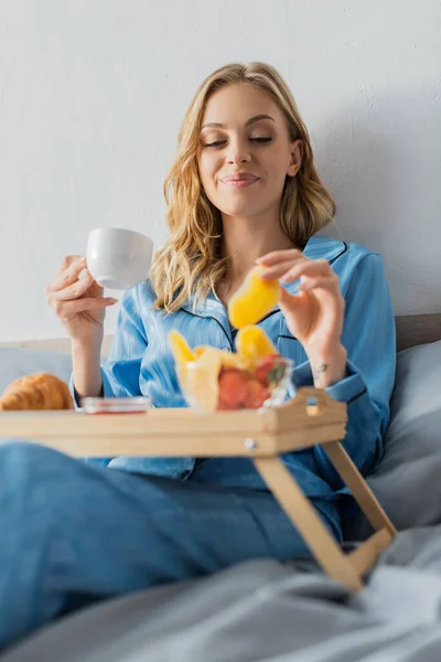 Sorrindo jovem mulher no pijama segurando xícara de café e manga seca perto bandeja enquanto toma café da manhã na cama — Fotografia de Stock