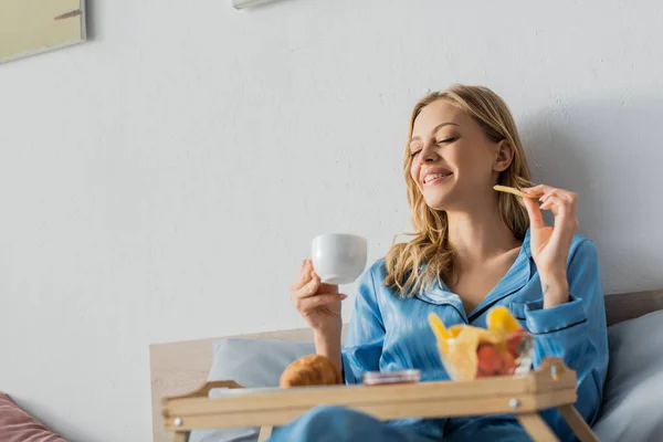 Joyful woman in pajama holding cup of coffee and dried mango near tray while having breakfast in bed — Stock Photo