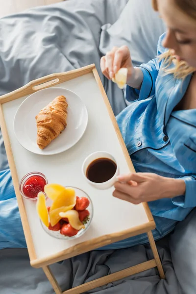 Vue de dessus de la femme tenant une tasse de café et croissant frais tout en prenant le petit déjeuner au lit — Photo de stock