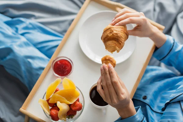 Vista superior da mulher cortada segurando croissant fresco perto da bandeja enquanto toma café da manhã na cama — Fotografia de Stock