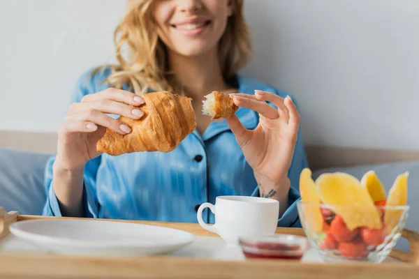 Vista cortada de sorrir jovem no pijama segurando croissant fresco perto da bandeja com café enquanto toma café da manhã na cama — Fotografia de Stock