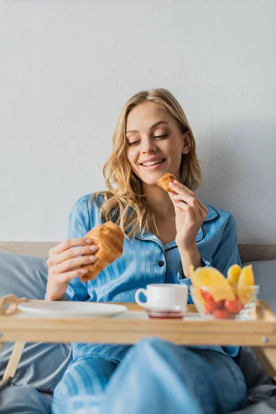 Mulher feliz no pijama comer croissant fresco perto da bandeja enquanto toma café da manhã na cama — Fotografia de Stock