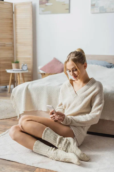 Jeune femme blonde en tricot pull et chaussettes bavarder sur smartphone dans la chambre moderne — Photo de stock