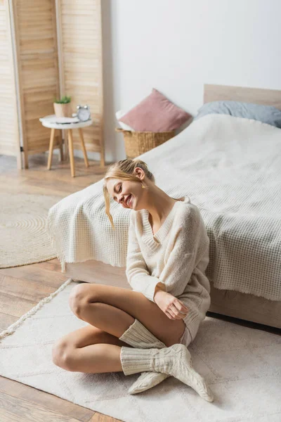 Mujer rubia joven en suéter de punto y calcetines sentados en la alfombra en el dormitorio moderno - foto de stock
