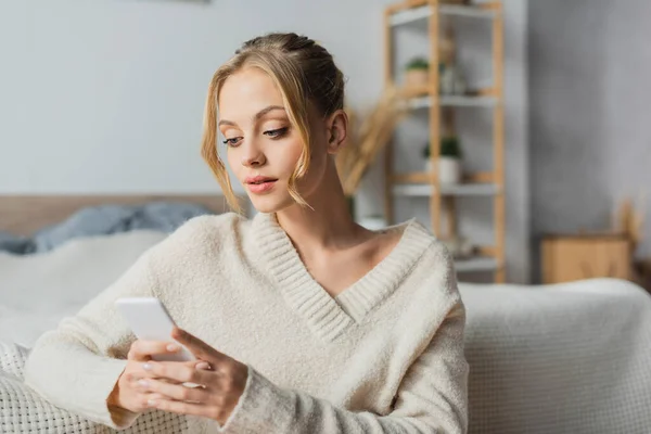Молода блондинка в в'язаному светрі спілкується на смартфоні в сучасній спальні — стокове фото