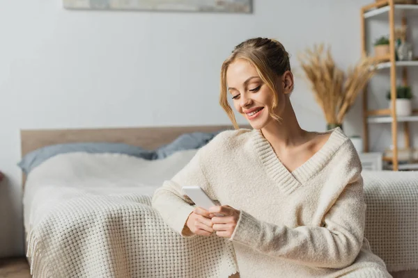 Allegra giovane donna in maglione utilizzando smartphone in camera da letto moderna — Foto stock