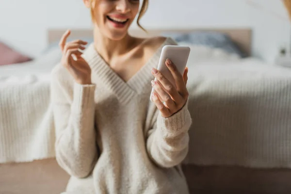 Vista recortada de la mujer feliz en jersey de punto usando teléfono inteligente en dormitorio moderno - foto de stock