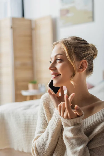 Allegra giovane donna in maglione che parla su smartphone in camera da letto moderna — Foto stock