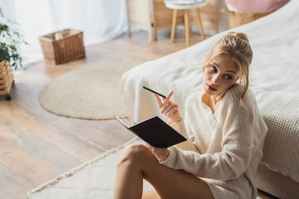Young blonde woman in sweater talking on smartphone and holding pen near notebook in bedroom — Stock Photo