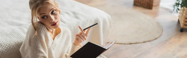 Blondine im Pullover spricht mit Smartphone und hält Stift neben Notizbuch im Schlafzimmer, Banner — Stockfoto