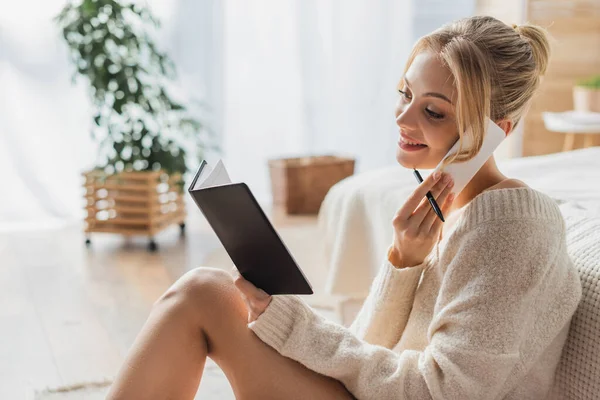 Fröhliche Frau in Pullover und Socken, die mit dem Smartphone spricht und aufs Notizbuch schaut — Stockfoto