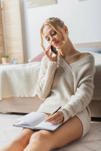 Mulher loira em suéter falando no smartphone e tomando notas no notebook — Fotografia de Stock