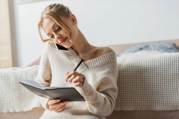 Hübsche Frau im Pullover, die mit dem Smartphone spricht und Notizen im Notizbuch macht — Stockfoto