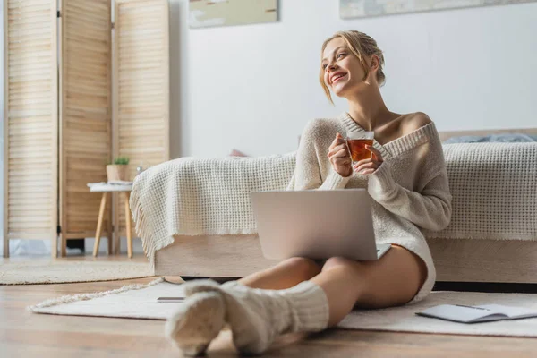 Щаслива жінка тримає скляну чашку чаю, сидячи з ноутбуком в сучасній квартирі — стокове фото