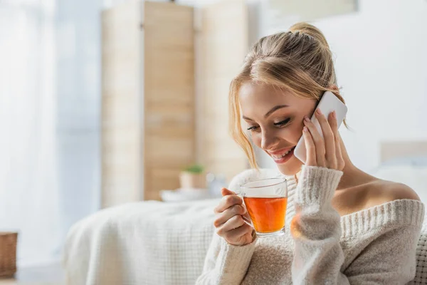 Mulher feliz segurando xícara de vidro de chá enquanto fala no smartphone no apartamento moderno — Fotografia de Stock